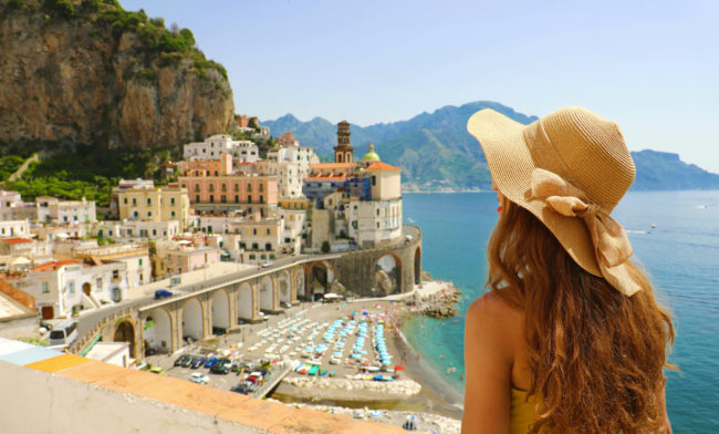 Summer holiday in Italy. Back view of young woman with straw hat and yellow dress with Atrani village on the background, Amalfi Coast, Italy